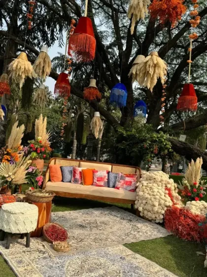 A bohemian-style Mehendi seating area under a large tree. The decor features hanging pampas grass and colorful lampshades, a rustic wooden sofa with vibrant cushions, and textured rugs on the grass.