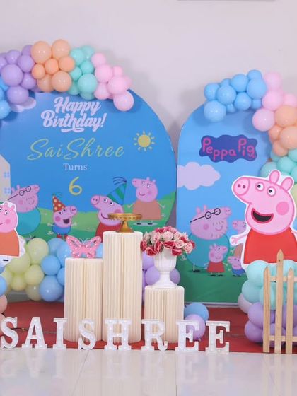 A fun and playful Peppa Pig themed birthday party. This setup includes two custom-arched backdrops with the Peppa Pig family, pastel balloon garlands, and a cute white picket fence.
