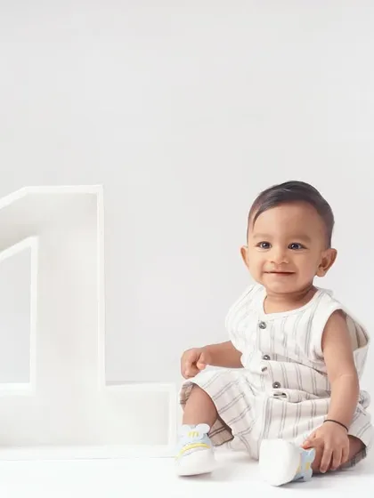 A happy baby boy in a striped romper smiles brightly while sitting next to a giant '1' for his minimalist first birthday shoot.