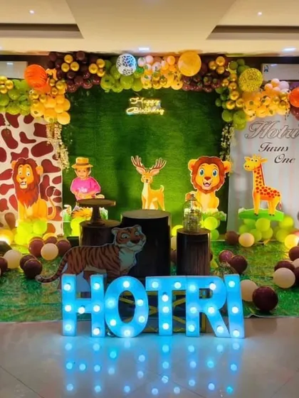 A cheerful jungle safari theme for a first birthday. The stage is decorated with a green foliage backdrop, cute cartoon animal cutouts, a light-up number '1', and a personalized name sign.