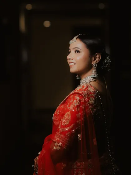 A candid shot of the bride, Divya, looking up with a hopeful smile. The lighting highlights her joy and the beautiful details of her red saree.