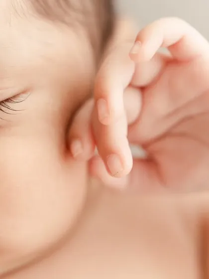 A close-up of a baby's hand resting gently on their face, showing their long eyelashes and soft skin.
