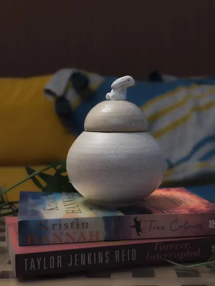 A serene, speckled white jar with a tiny rabbit on the lid, resting on a stack of books. This piece combines functionality with a subtle, charming design.