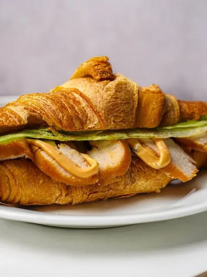 A side profile of a chicken-filled croissant sandwich, showing the layers of ingredients. The bright, clean aesthetic is ideal for online menus and promotional materials.