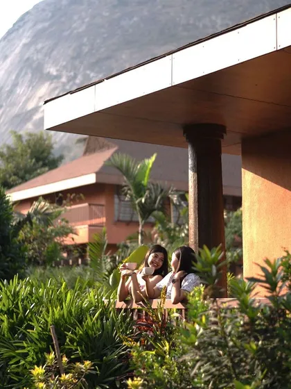 Two guests share a conversation on a bungalow porch, with the majestic Nandi Hills in the background. Our accommodations are designed to offer both privacy and stunning views.