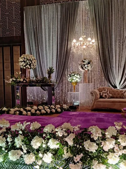 A sophisticated stage for a Sagan or Sangeet ceremony. The decor features shimmering silver and grey drapes, a plush vintage sofa, and an abundance of white flowers for a classic and luxurious feel.
