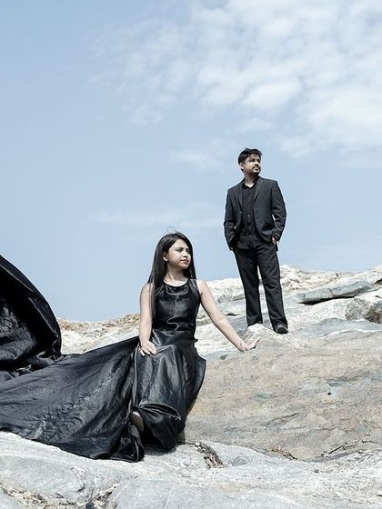 A couple stands on a rocky cliff, her black trail gown billowing against the sky, creating a dramatic and powerful pre-wedding portrait.