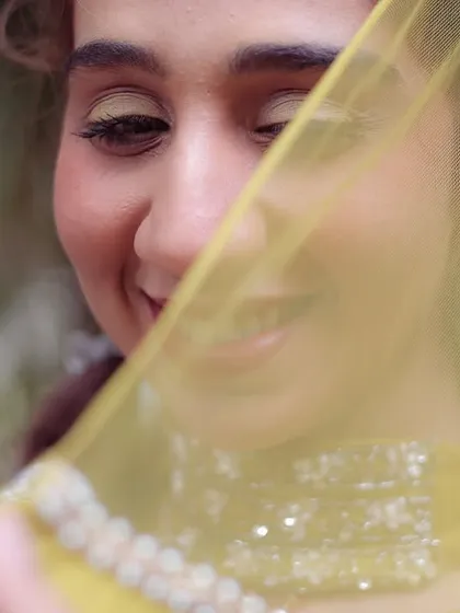 A close-up portrait of a bride, her face partially veiled by her yellow dupatta. This shot from a Haldi or Mehendi ceremony is full of warmth and shy happiness.