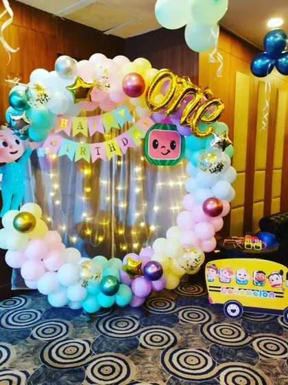 A Cocomelon-themed ring backdrop with fairy lights, character cutouts, and a 'ONE' foil balloon for a first birthday.