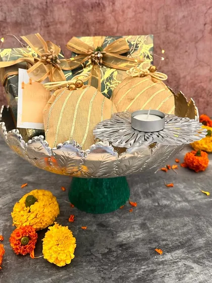 A festive hamper arranged in a silver-finish urli with a green velvet base. It includes beautifully wrapped gift boxes and a matching T-light holder, creating a rich and traditional look.