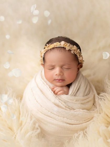 Pure and serene. This baby is wrapped in a soft cream swaddle, nestled on a fluffy blanket with delicate white petals falling around her.
