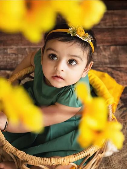 An artistic shot of a baby girl in a basket, viewed through a foreground of yellow flowers.