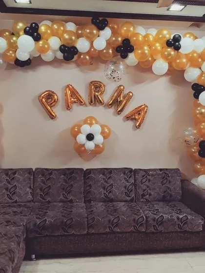 A home birthday decoration with a gold, white, and black balloon arch over the sofa. The person's name is spelled out in gold foil letters in the center of the arch.