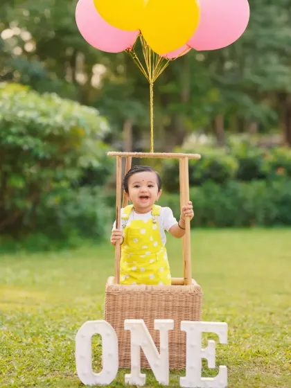 A bright and cheerful first birthday photo with a hot-air balloon prop and colorful balloons, perfect for a sunny day in the park.