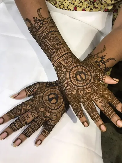 A special mehendi design for a baby shower, featuring tiny baby feet inside a circle and the words 'Mom to be' and 'Dad to be' on the fingers.