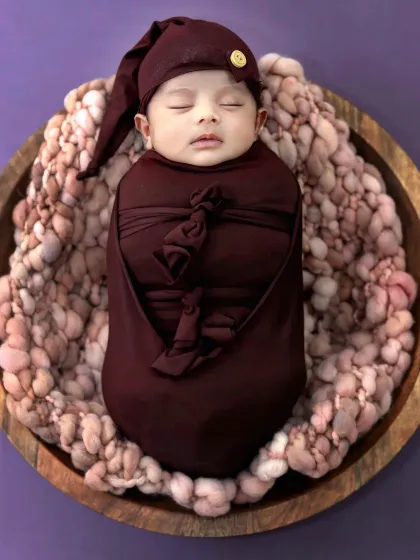 A perfectly swaddled newborn sleeps in a wooden bowl, looking cozy and content in a deep maroon wrap.