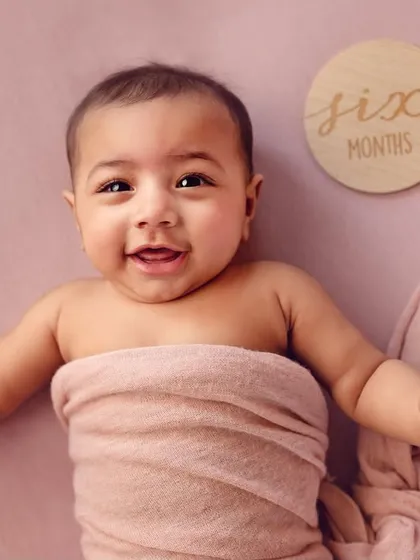 A happy six-month-old baby wrapped in a pink cloth. Milestone sessions for 4 or 6-month-olds are perfect for capturing their developing personalities and adorable expressions.