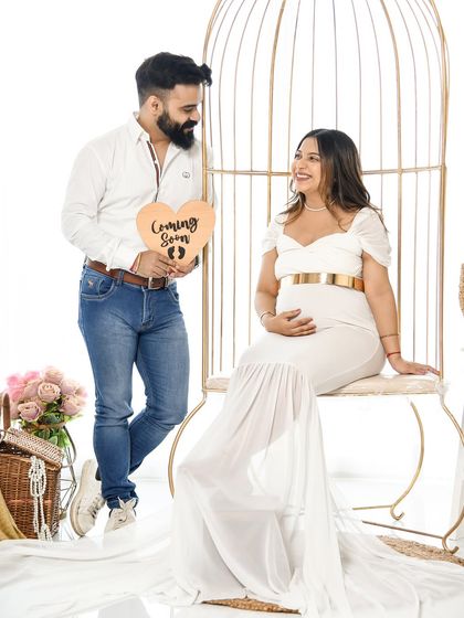 A joyful couple's portrait in our boho birdcage setup. The dad-to-be holds a "Coming Soon" sign, sharing a happy look with his partner.