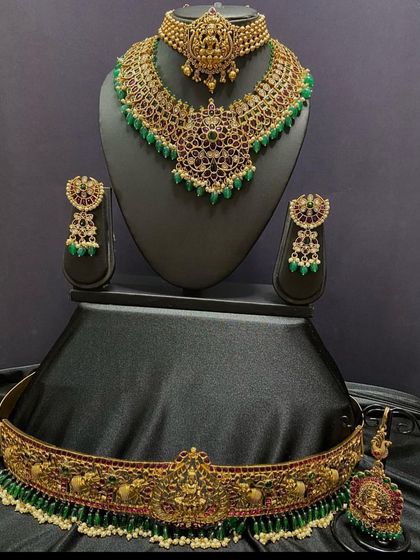 A complete bridal set with a kemp stone choker and a matching short necklace. The set also includes a vaddanam and maang tikka with green and red stone details.