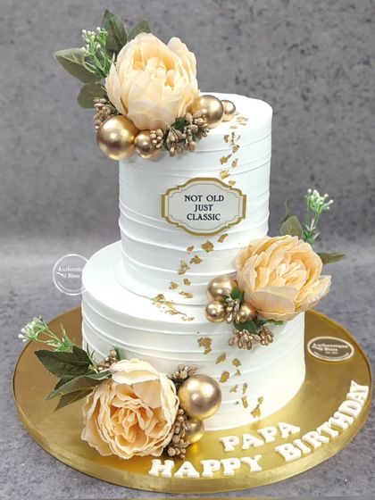 A classic two-tiered floral cake for a father's birthday. Adorned with peach-colored peonies and gold accents, the cake carries a tag that reads "Not Old Just Classic," a perfect sentiment for a milestone celebration.