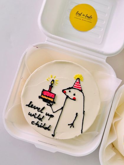 A quirky "level up wild child" bento cake with a hand-drawn character holding a mini cake and candle, perfect for a gamer or anyone celebrating a new chapter.