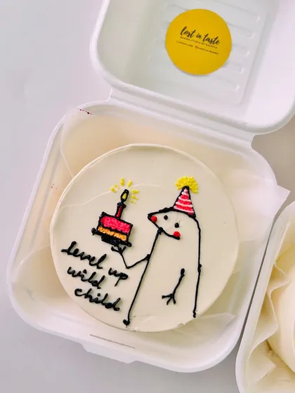 A quirky "level up wild child" bento cake with a hand-drawn character holding a mini cake and candle, perfect for a gamer or anyone celebrating a new chapter.