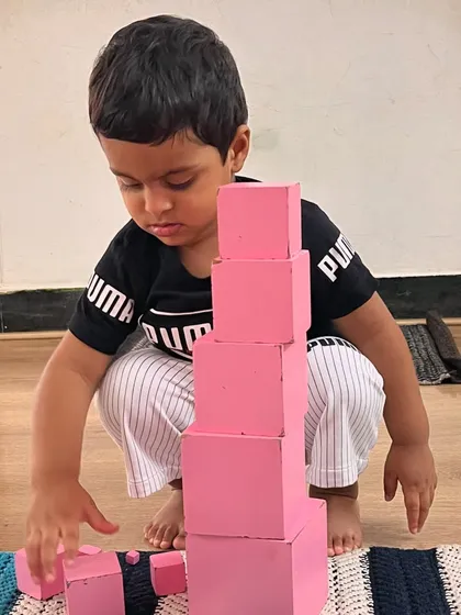 A young boy builds the Pink Tower, a classic sensorial material. This activity helps him visually discriminate dimensions and develop a sense of order and sequence from largest to smallest.