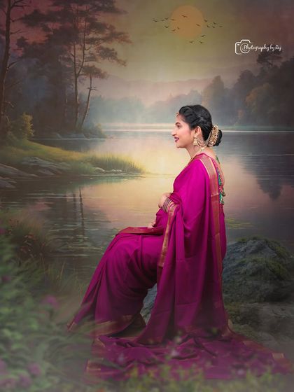 A beautiful studio portrait of a mother-to-be in a magenta saree, seated against a painted landscape backdrop.