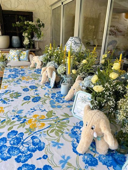 A long table decorated with a blue and yellow floral tablecloth, plush elephant toys, and arrangements of baby's breath.
