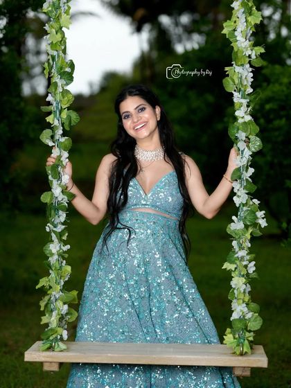 A joyful portrait of a mother-to-be in a sparkling blue two-piece lehenga, seated on our floral swing.
