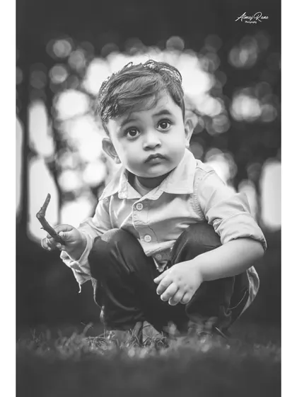 A pensive black and white portrait of Ivaan crouching in the grass. This image has a classic, timeless feel, focusing on his innocent and curious expression.