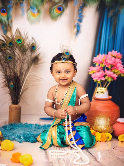 A classic portrait of a baby dressed as Krishna, sitting peacefully with his pearl necklace. The setup is complete with peacock feathers and marigolds.
