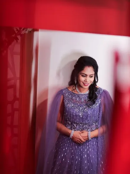 A candid portrait of a bride, framed by vibrant red curtains. Her shy smile and elegant purple gown create a beautiful, colorful image from her wedding day.