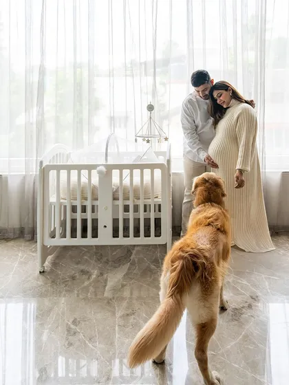 A serene and bright maternity photo. Hugo the Golden Retriever watches as his parents-to-be share a moment by the baby's crib, anticipating the new arrival.
