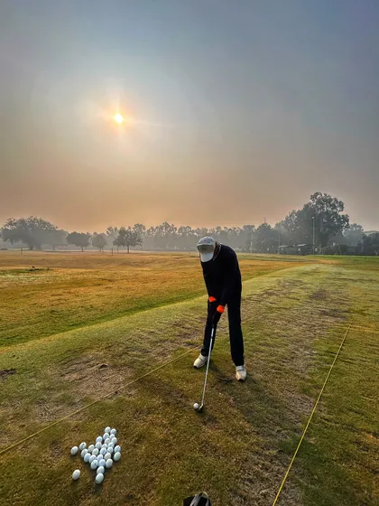 A golfer practices on the grass tee during the golden hour, with a full bucket of balls ready to go.
