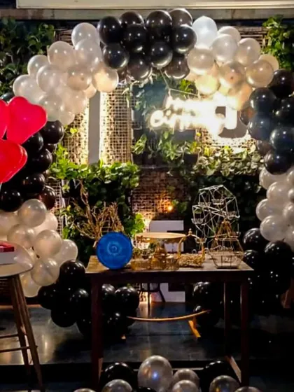 A sophisticated party setup with a black, white, and silver balloon arch, accented with red heart balloons for a pop of color. The decor is arranged around a rustic table against an exposed brick wall, perfect for a trendy cafe or restaurant celebration.