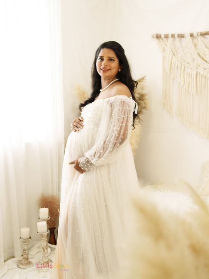 A radiant mother-to-be in a delicate white lace gown. She stands by a window in a bright boho room, the natural light creating a soft and beautiful glow.