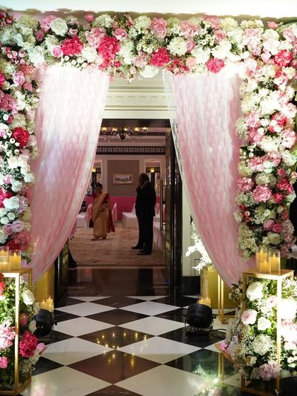 This entrance is a vision in pink and white. A dense floral archway with soft pink draping creates a fairytale-like passage, perfect for a romantic wedding theme, set against the classic checkered floor of the ITC Maratha.