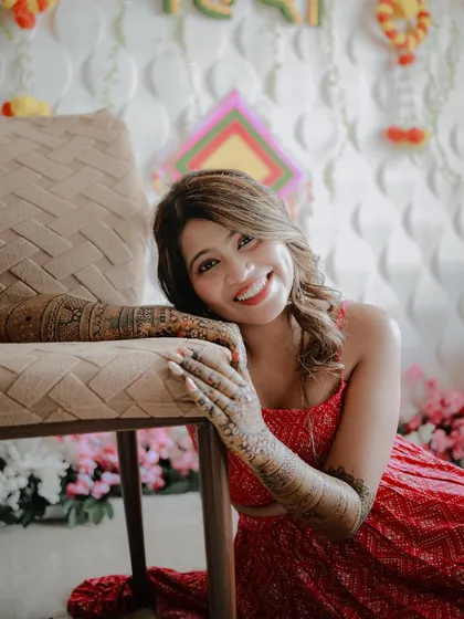 A bright and happy portrait of the bride. She rests her henna-covered arms on a chair, looking directly at the camera with a joyful and welcoming smile.