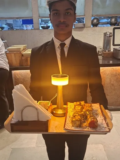 Our team ensures that every dish is presented perfectly. A server holds a tray with a beautifully plated appetizer and a warm table lamp, ready to offer guests a taste of our culinary artistry.