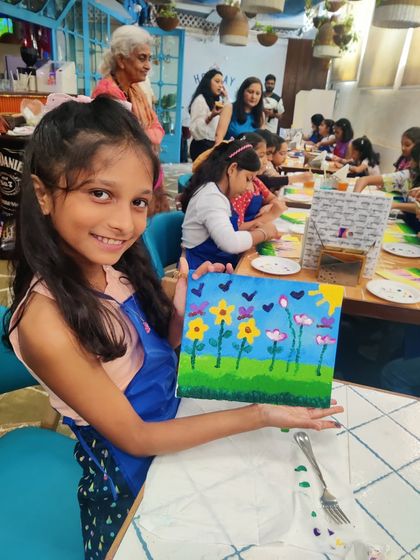 A young artist in a blue apron proudly displays her finished canvas at a birthday party held in a cafe. Her painting features a colourful garden with a bright sun, capturing the joyful atmosphere of the event.