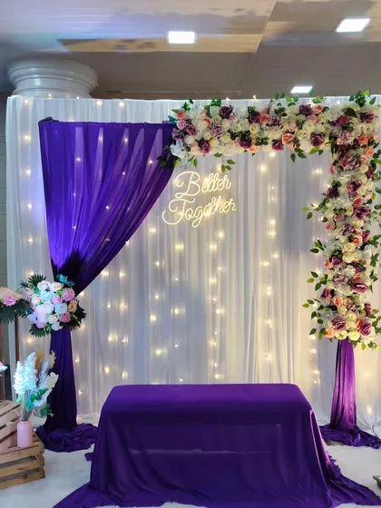 An intimate engagement photo corner featuring a rich purple and white color scheme. We used elegant draping, a backdrop of fairy lights, and an asymmetrical floral arch, completed with a custom "Better Together" neon sign for a modern, romantic touch.