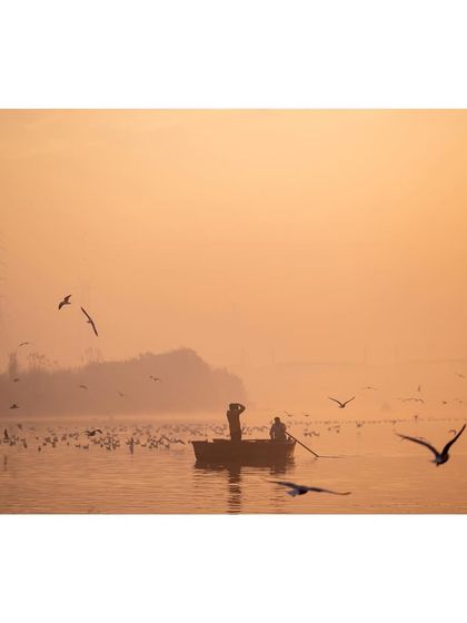 A boatman and passenger on the Yamuna River at sunrise, surrounded by a flock of migratory birds. The hazy, golden light creates a dreamy and picturesque scene.