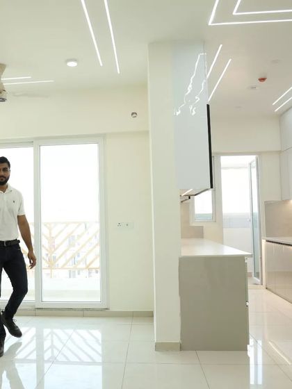 A wide shot of a living room with a high ceiling and large windows. The space is ready for furniture, with the false ceiling, profile lighting, and wall lights already installed.