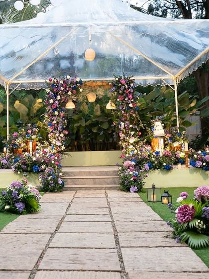 A fairytale wedding reception under a clear-top tent. The aisle is lined with candle-lit lanterns, leading to a stage overflowing with whimsical purple and blue flowers.