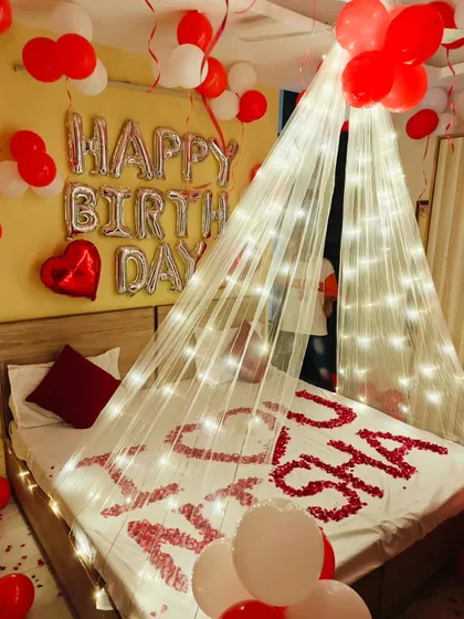 A personalized birthday message, "I ❤️ U NISHA," is spelled out with rose petals on the bed. The scene is framed by a light-up canopy and red and white balloons.