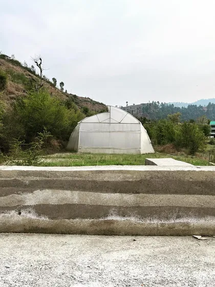 A finished rammed earth wall stands in the foreground of the Himalayan landscape. In the background, a polyhouse for farming shows the connection between our architectural work and the agricultural context of the region.