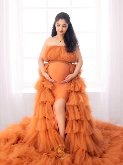 A classic solo maternity portrait against a bright window. The mother-to-be looks down lovingly at her bump, dressed in a beautiful tiered orange gown.