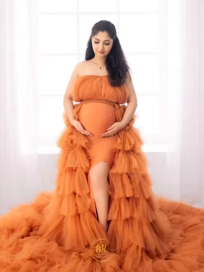 A classic solo maternity portrait against a bright window. The mother-to-be looks down lovingly at her bump, dressed in a beautiful tiered orange gown.
