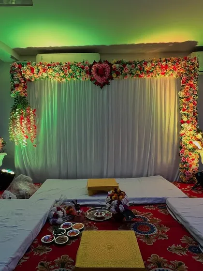A simple mandap setup for a home or small hall. The backdrop is a white curtain with a floral arch, and the seating is arranged on the floor.